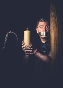 Author photo of alarmed looking Darren Shan in a darkened room, duck tape over his mouth and bound hands holding a candle, with a sinister young girl lurking in the background